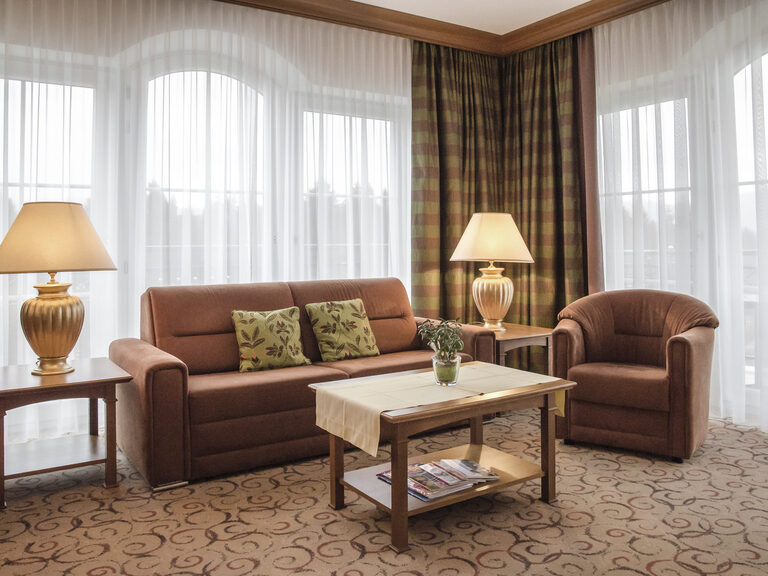Der luxuriöse Wohnbereich der Turmsuite Kronberg im Wellnesshotel Mooshof in Bodenmais bietet einen komfortablen Sitzbereich mit einen eleganten Couchtisch vor den deckenhohen Fenstern.