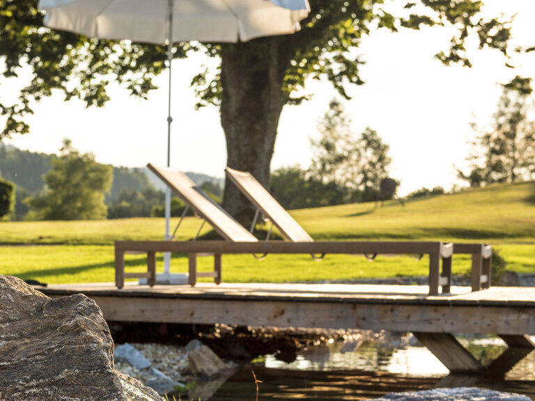 Die idylische Uferlandschaft am Naturbadesee des Wellnesshotels Mooshof in Bodenmais mit gemütlichen Liegen und Sonnenschirm.