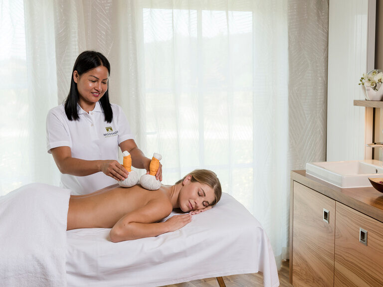 Im Behandlungsraum des Vitalresorts des Wellnesshotels Mooshof in Bayern erhält eine Frau eine entspannende Rückenmassage auf einem Massagebett.
