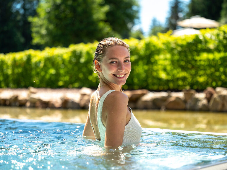 Eine Frau badet glücklich im strahlend blauen Wasser des Erholungspools des Wellnesshotels Mooshof im bayerischen Wald.