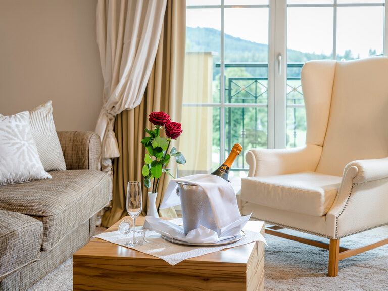 Der wohnliche Raum mit Champagner als Begrüßungsgeschenk und gemütlicher Sitzecke vor einem großen Fenster in der Honeymoonsuite des Wellnesshotels Mooshof im Bayerischen Wald.