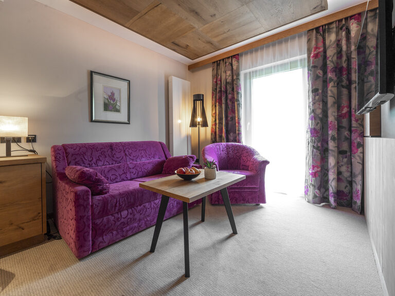 Das Zimmer Typ 1 des Wellnesshotels Mooshof in Bodenmais empfängt Gäste mit einem stilvollen lila Sofa im Wohnzimmer.