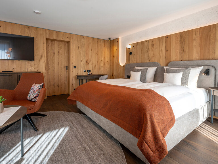 Einladendes Bett im Wellnesshotel Mooshof im Bayerischen Wald, neben einem gemütlichen Stuhl im Zimmer Typ 3, das durch seinen modernen Fernseher besticht.